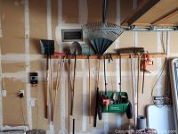 Full view of the gardener tool set hanging on a garage wall including shovels, rakes, broom, electric soil auger, spreader bucket with handheld tools inside, and apron.
