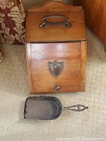 Front view of wooden coal bin with metal handle on lid, shield-shaped front vent, and metal shovel on floor.