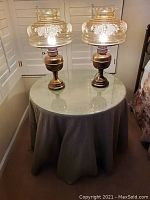 Full view of the bedside table with glass top and cloth skirt holding two antique lamps with etched glass globes.