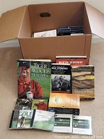 View of golf instruction books and media laid out in front of a cardboard box showing titles and DVD cases.