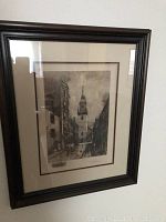 Framed black and white urban scene artwork by Robert Shaw, with church steeple and street with people, in dark wooden frame