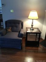Blue armchair next to wooden side table with glass top and brass lamp with shade