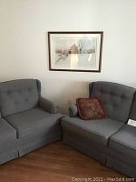 Photo showing two blue loveseats positioned at right angles with a red decorative pillow and a framed print hung on the wall above them.