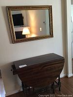 Solid wood drop leaf table shut with framed gold leaf mirror mounted above it on the wall.