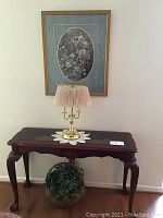 Photo of a wooden sofa table with a brass touch lamp on top, a framed floral artwork hanging on the wall behind, and a round woven basket with greenery underneath the table.