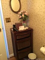 Photo of wooden shelf unit with 3 woven wicker baskets in a corner below an oval wall mirror mounted on wallpapered wall. Shelf top contains decorative box, lace doily and black boot-shaped decoration.