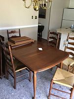 Dining table with 3 leaves stacked on top and 7 ladder back chairs with cane seat arranged around the table in dining room setting.