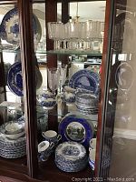 Photo showing multiple shelves of blue and white Liberty Blue china including plates, bowls, cups, and saucers along with clear glassware including water and wine glasses in dark wood cabinet.