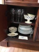 Open cabinet shelf showing stacked china plates, small plates, teacups, and glass goblets.