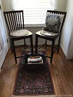 Two wooden bar stools with vertical slat backs, seat cushions, patterned rug beneath, basket with books between them, pillow on one stool.