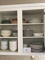 Cabinet with multiple shelves showing various bowls and plates stacked neatly including glass, ceramic and woven baskets.