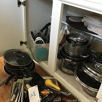 View of cabinet with multiple stacked cooking pots and pans with lids, plastic container with kitchen utensils, and cabinet shelves with folded kitchen towels and mats.