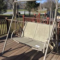 Beige metal framed outdoor porch swing with mesh seating on wooden porch deck, showing weathered use.