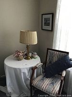 Accent chair, round table covered with white cloth, brass table lamp, and framed artwork on wall beside window.
