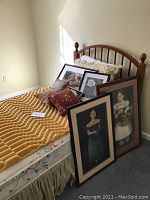 View of twin bed with wooden headboard, mattress with yellow topper, and four framed artwork pieces leaning on bed, plus two pillows