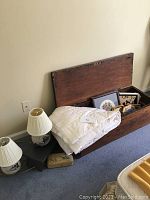 Barnwood trunk open, containing framed artwork and mirrors, white down quilt folded over the edge, two pottery lamps with white pleated shades on floor nearby, small embroidered pillow on floor.
