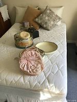 Double bed with white quilted mattress. Several vintage hats and hat boxes on the mattress with pillows behind.