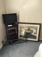 Small Toshiba flat screen TV on top of a dark brown wooden bookshelf with three shelves. Next to the bookshelf is a framed artwork resting on the floor and a metal fireplace screen with a geometric pattern also on the floor.