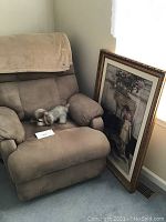 Tan recliner with a small plush dog toy on the seat and a beige throw draped over the backrest. The recliner is placed against a wall near a window.
