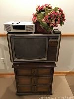 Photo showing the Bose Wave Radio stereo remote, vintage wood-paneled television sitting on the small wooden dresser, and artificial flowers in a basket on top of television