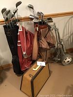 Photo showing two sets of vintage golf clubs in aged black and reddish-brown bags, a vintage tan hard travel case, and a foldable metal golf cart.