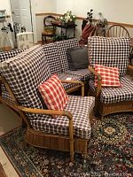Front angle of three-piece rattan couch set with blue and white plaid cushions and red plaid pillows on armchairs, positioned on patterned rug in a room with various furniture and storage items.