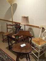 Photo of two bar-height chairs (one white with woven seat, one brown wooden swivel chair) and one standard height wooden chair beside a wooden side table with table lamp and floor lamp behind it.