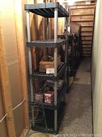 Four black heavy duty plastic shelving units in a narrow wooden room with various items on the shelves including holiday decor, painting supplies, baskets, and wire frames