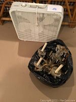 White plastic box fan with adjustable speed dial and pull cord standing upright against wall on floor, next to black trash bag containing chandelier parts.