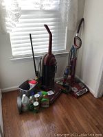 Photo showing two upright vacuum cleaners (Dirt Devil Maxima and Shark) and several miscellaneous cleaning supplies arranged on hardwood floor by window.