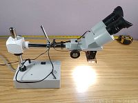 Side angle of stereo microscope showing dual eyepieces, illumination light on, and universal adjustable stand with base and pole.
