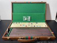 Domino set with ivory-colored tiles numbered in black, red, and blue arranged inside an open wood grain case lined with green felt and brown fabric, including two dice in a small plastic bag.