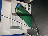 Photo showing boxed mini putting green golf mat, metal putter, and multiple golf balls arranged on the box and mat surface. The mat depicts a green putting surface with a sand trap and water hazard, as well as holes for putting.