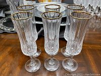 Photo showing six crystal champagne flutes with 24k gold rim placed on a wooden surface, with a silver tray in the background.