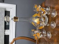 Side view of the glass decanter with stopper and six matching stemmed glasses on a wooden table, showing the orange rose floral pattern and gold accents.