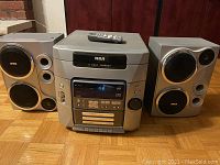 Front view of the RCA stereo system showing the main unit with control panel, digital display, 5 disc CD changer on top, and two matching speakers on each side with RCA logos.