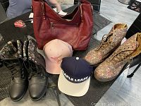 Full lot display showing red leather handbag, black combat-style Doc Martens boots with floral lining, floral patterned lace-up boots, pink Toronto Blue Jays cap, and navy beige Toronto Maple Leafs cap on glass table.