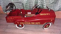 Side view showing red metal body with 'VOLUNTEER FIRE DEPT.' and 'TRUCK No 1' lettering, whitewall tires, and black pedals and seat.