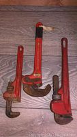 Three pipe wrenches laid out side by side on a floor: a 10 inch adjustable wrench, a 12 inch clamp-type wrench, and a 14 inch adjustable wrench, all red with visible signs of use and rust.