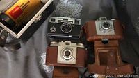 Three vintage Kodak cameras laid out on fabric surface with two in leather cases and one loose.