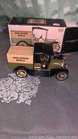 Photo showing the side and front of Harley Davidson 1916 Studebaker pickup model bank next to its box, details of green, black and gold coloring.