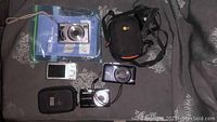 Four small digital cameras with two camera cases, charger plug, and instruction booklet arranged on a patterned surface.