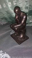 Bronze-colored statuette of a muscular male figure seated in a thinking pose, shown at an angle to highlight side profile and base.