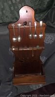 Front view of wooden wall-mounted display rack holding miniature souvenir spoons and forks with a pull-out bottom drawer.