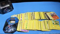 Loose assorted Pokemon trading cards spread out next to the Pokemon tin with Greninja artwork visible on lid. Cards are a mix of types and sets.