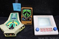 Photo showing all three handheld video games together: Coleco beige device shaped with baseball diamond, Mattel brown rectangular with green baseball diamond, Ohio Art white device with screen and red buttons with blue control knobs.