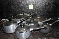 Full set of pots and pans arranged on a flat surface, showing variety of sizes and presence of lids, some with glass tops and black handles