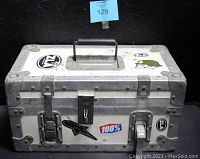 Closed metal lockable road case box with stickers on exterior, including black and white logos, a green character, and a '100%' sticker. Top handle visible.