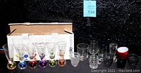 Box with 6 clear vodka shooter shot glasses with colorful bases arranged in front of box labeled Bonfire Circle Ware. Additional 10 assorted decorative vintage shot glasses placed nearby with various designs and colors.