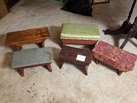 Five small antique footstools and step stools grouped on carpeted floor showing variety of finishes and upholstery conditions.